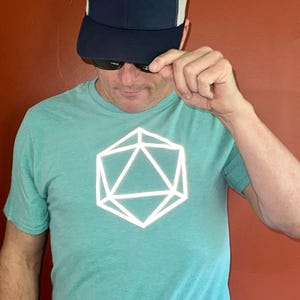 May include: A light teal t-shirt featuring a white geometric design. The design is a stylized 3D shape. The person in the image is wearing a navy blue and white trucker hat and sunglasses.