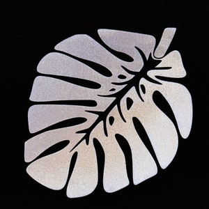 Pode incluir: Um recorte de folha de monstera de glitter prateado, uma planta tropical popular, sobre um fundo preto.