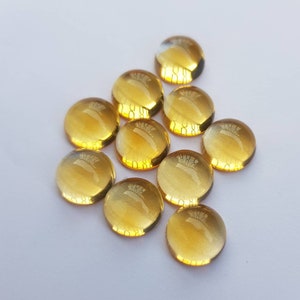 May include: A group of ten round, translucent, yellow glass cabochons. The cabochons are arranged in a loose, triangular pattern on a white surface.