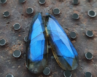 May include: Two teardrop-shaped blue labradorite gemstones with iridescent shimmer on a rusty metal background.