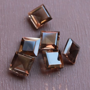 May include: Six brown square-shaped gemstones with a faceted cut. The gemstones are arranged in a cluster on a brown surface.