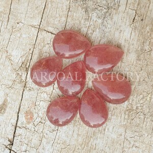 May include: Seven teardrop-shaped, translucent pink gemstone cabochons. The stones have a slightly rough texture and are arranged in a cluster on a wooden surface.