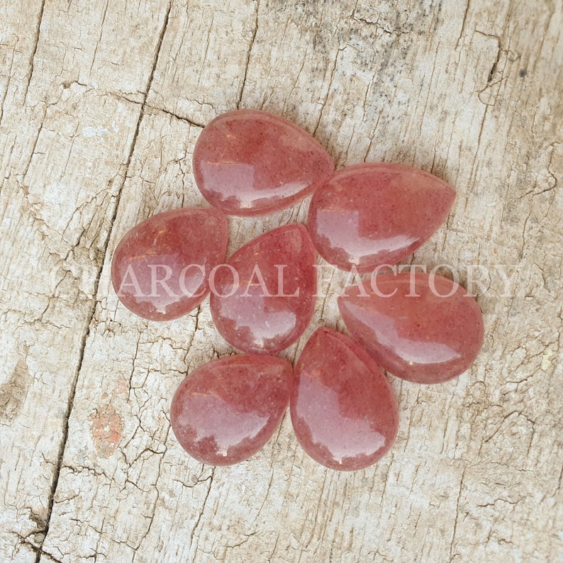 May include: Seven teardrop-shaped, translucent pink gemstone cabochons. The stones have a slightly rough texture and are arranged in a cluster on a wooden surface.