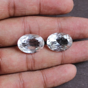 May include: Two clear oval-shaped faceted gemstones, likely quartz, displayed on a hand.
