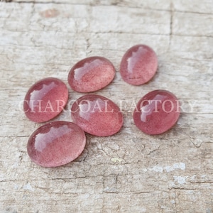 May include: Six oval-shaped, translucent pink gemstones with a slightly cloudy appearance. The stones are arranged on a light brown wooden surface.