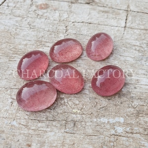 May include: Six oval-shaped, translucent pink gemstones with a sparkly, speckled texture. The stones are arranged on a light brown wooden surface.