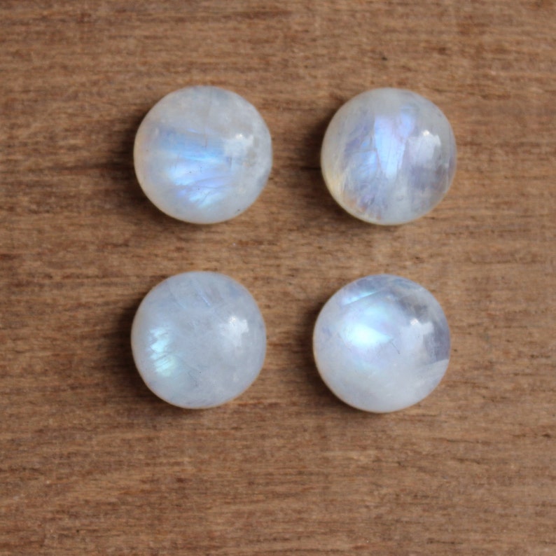 May include: Four round, white moonstone cabochons with iridescent blue and white flashes. The stones are arranged in a square pattern on a wooden surface.