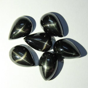 May include: Six black star sapphire gemstones, teardrop-shaped, are arranged on a white surface. Each polished stone displays a star effect, with a cross-shaped light reflection across the surface.