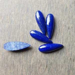 Puede incluir: Cinco piedras de lapislázuli en forma de lágrima de diferentes tamaños, con una sexta piedra de lapislázuli plana, más pequeña. Las piedras son de un azul profundo con destellos dorados.