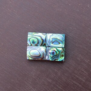 May include: Four square tiles made of iridescent abalone shell. The tiles are arranged in a 2x2 grid. The tiles have a green, blue, and purple color scheme.