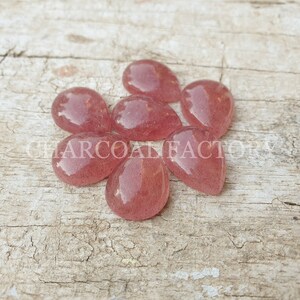 May include: Seven pink teardrop-shaped gemstone cabochons, each with a slightly different shade of pink, are arranged on a light brown wooden surface.