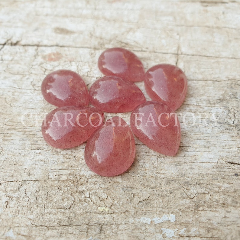 May include: Seven pink teardrop-shaped gemstone cabochons, each with a slightly different shade of pink, are arranged on a light brown wooden surface.