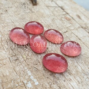 May include: Six oval-shaped, translucent pink gemstones with a speckled texture. The gemstones are arranged in a cluster on a wooden surface.