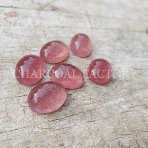 May include: Seven oval-shaped, translucent pink stones with a speckled texture. The stones are arranged on a light brown wooden surface.