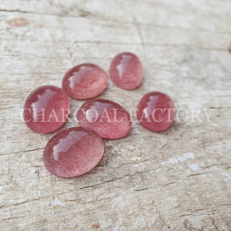 May include: Seven oval-shaped, translucent pink stones with a speckled texture. The stones are arranged on a light brown wooden surface.