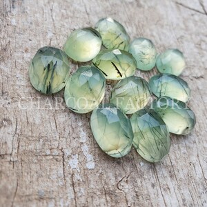 Op de afbeelding: Een verzameling van 12 groene en zwarte edelsteen cabochons met een gefacetteerde ovale vorm. De stenen hebben een doorschijnende uitstraling met zwarte insluitsels.