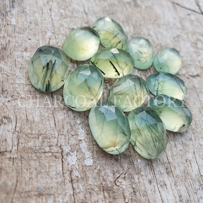 Op de afbeelding: Een verzameling van 12 groene en zwarte edelsteen cabochons met een gefacetteerde ovale vorm. De stenen hebben een doorschijnende uitstraling met zwarte insluitsels.