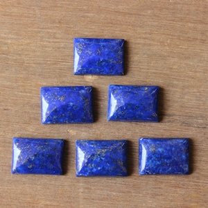 May include: Six rectangular lapis lazuli gemstones, each with a deep blue hue and gold inclusions, are displayed on a wooden surface. The polished stones have a glossy finish, reflecting light.