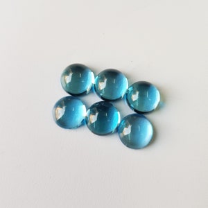 Op de afbeelding: Zes ronde, blauwe topaas edelstenen. De edelstenen zijn glad en hebben een licht iriserende glans.