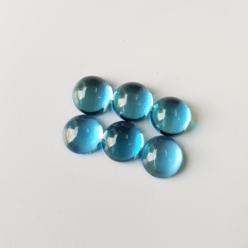 Op de afbeelding: Zes ronde, blauwe topaas edelstenen. De edelstenen zijn glad en hebben een licht iriserende glans.