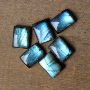May include: Six iridescent blue-green labradorite gemstone cabochons, cut into rectangular shapes, are arranged on a wooden surface.