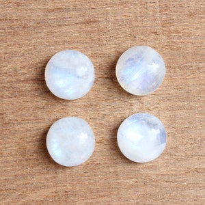May include: Four round, white moonstone cabochons with iridescent blue and white flashes. The stones are arranged in a square pattern on a brown wooden surface.