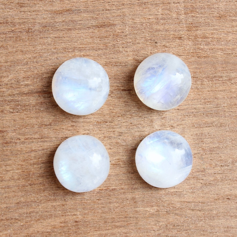 May include: Four round, white moonstone cabochons with iridescent blue and white flashes. The stones are arranged in a square pattern on a brown wooden surface.