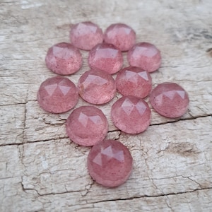 May include: A collection of 12 faceted, pink gemstone cabochons. The cabochons are arranged in a semi-circle pattern on a wooden surface.