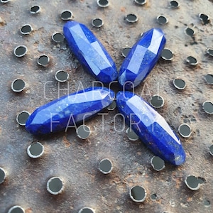 Pode incluir: Quatro pedras preciosas de lapis-lazúli facetadas, de forma oval, de cor azul escura. As pedras estão dispostas em forma de estrela sobre uma superfície metálica enferrujada.