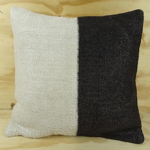 May include: Square cushion with a two-tone design. One half is a light beige colour, and the other half is a dark charcoal grey. The cushion has a textured, woven appearance, suggesting a natural fibre material.