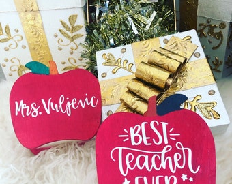 May include: Two red apple-shaped wooden signs with white lettering. One sign says "Mrs. Vuljevic" and the other says "Best Teacher Ever".