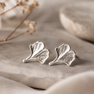 Op de afbeelding: Een paar zilveren oorknopjes in de vorm van ginkgobladeren. De oorbellen hebben een getextureerd, metalen oppervlak met een opvallend waaiervormig silhouet en een steeltje, wat de organische vorm van het blad in zilveren afwerking weergeeft.