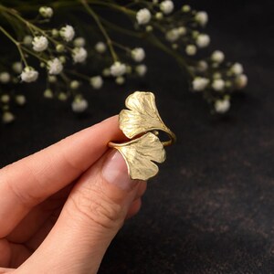 May include: A gold-coloured ring featuring a ginkgo leaf design. The ring has two stylised leaves that wrap around the finger. The leaves have detailed veining and a textured surface. The ring is shown against a dark background with white flowers.