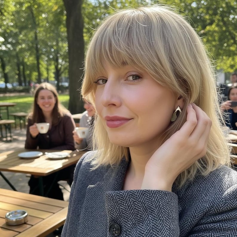 May include: A gold-toned metal earring with a curved, organic shape worn on the earlobe. The earring features a smooth, polished surface that reflects light, creating a subtle contrast against the skin and blonde hair of the wearer.