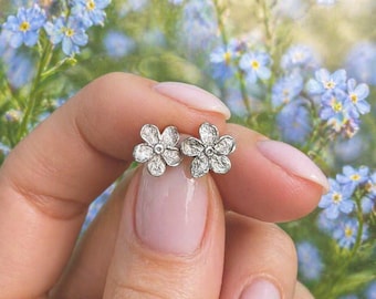 Puede incluir: Un par de pendientes de botón plateados con forma de flores de cinco pétalos y un acabado metálico texturizado. Los pendientes presentan un punto central y pétalos delicados y redondeados, con un diseño detallado y uniforme.