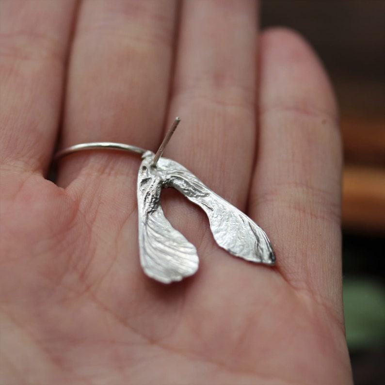 May include: A silver stud earring shaped like a maple seed, also known as a samara. The metallic piece features a textured, organic surface with a thin post extending from the top, capturing the distinct dual-wing structure of a botanical seed.