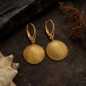 Könnte beinhalten: Ein Paar goldfarbene Ohrringe mit Muscheldesign. Die Ohrringe haben einen runden, fächerförmigen Anhänger mit detaillierten Linien und einen Klappverschluss. Die Ohrringe werden auf einer dunklen, strukturierten Oberfläche präsentiert.