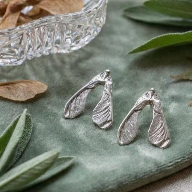 May include: A pair of silver-toned stud earrings shaped like maple tree samara seeds. Each earring features a textured, organic surface with a bifurcated wing design that mimics the natural appearance of a winged seed pod in a metallic finish.