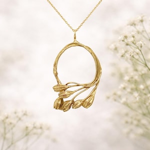 May include: A gold-colored pendant necklace featuring a circular design with a floral motif. The pendant has a textured, organic appearance, with a delicate chain. The necklace is set against a soft, blurred background.