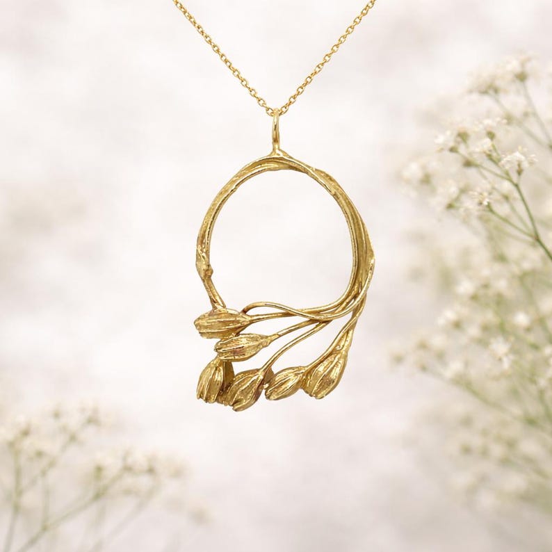 May include: A gold-colored pendant necklace featuring a circular design with a floral motif. The pendant has a textured, organic appearance, with a delicate chain. The necklace is set against a soft, blurred background.