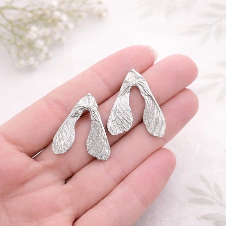 May include: Two silver-toned metal objects shaped like maple tree samara seeds, also known as helicopter seeds. Each piece features a textured, veined surface mimicking the organic structure of a winged seed pod with a V-shaped connection point.
