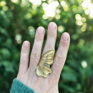 Op de afbeelding: Een gouden vlinder ring met een getextureerd oppervlak. De ring wordt gedragen op de middelvinger van een hand.