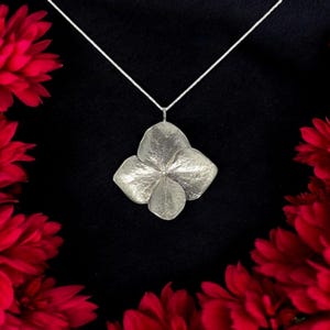 Peut inclure: Un collier pendentif argenté avec un motif de fleur à quatre pétales. Le pendentif est suspendu à une fine chaîne argentée. L'image est posée sur un fond noir, avec des fleurs rouges dans les coins.