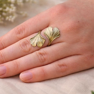 Könnte beinhalten: Ein goldfarbener Ring mit einem Ginkgoblatt-Design. Der Ring hat zwei blattförmige Elemente, die sich um den Finger legen. Die Blätter haben detaillierte Adern und eine strukturierte Oberfläche, wodurch ein naturalistisches Aussehen entsteht. Der Ring wird an einer Hand getragen.