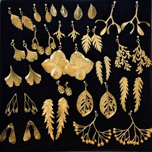 May include: A collection of gold-colored leaf-shaped earrings, displayed on a black surface. The earrings vary in design, including fern, ginkgo, and other botanical forms. Some earrings feature intricate details, while others are simpler in design. The earrings are presented in a grid-like arrangement.