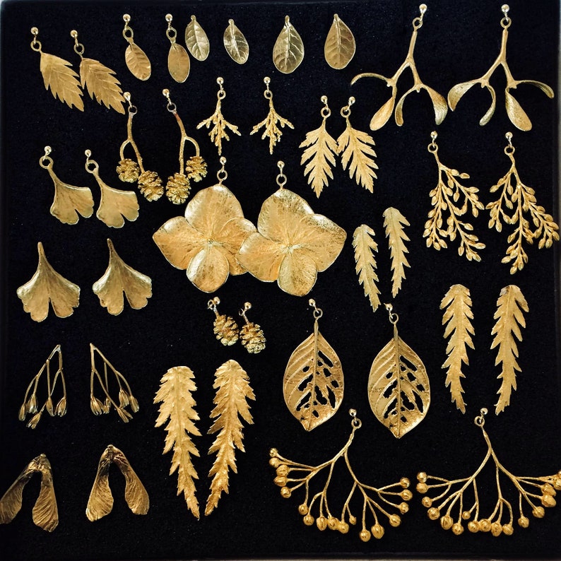 May include: A collection of gold-colored leaf-shaped earrings, displayed on a black surface. The earrings vary in design, including fern, ginkgo, and other botanical forms. Some earrings feature intricate details, while others are simpler in design. The earrings are presented in a grid-like arrangement.