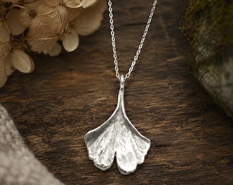 Puede incluir: Un collar de tono plateado con un colgante en forma de hoja de ginkgo. El colgante tiene una superficie texturizada y orgánica con venas visibles y un acabado metálico. Cuelga de una cadena fina de metal de tono plateado con eslabones.