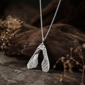 May include: A silver-colored pendant shaped like a maple seed pod, suspended from a delicate matching silver-colored chain. The pendant features a textured, organic surface that mimics the veins and structure of a natural winged seed.