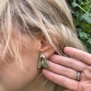 May include: A silver-toned earring shaped like a maple seed pod, also known as a samara, featuring a textured surface and a bifurcated design. The earring is positioned on the earlobe, showcasing its organic, wing-like structure and metallic finish.