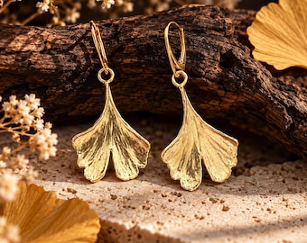 May include: A pair of gold-toned drop earrings featuring a textured ginkgo leaf design. Each earring is suspended from a simple gold-toned leverback closure, showcasing a detailed, fan-shaped leaf with visible veins and a metallic finish.